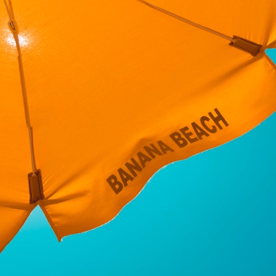 An orange beach umbrella against a blue sky, with the words Banana Beach written on the umbrella